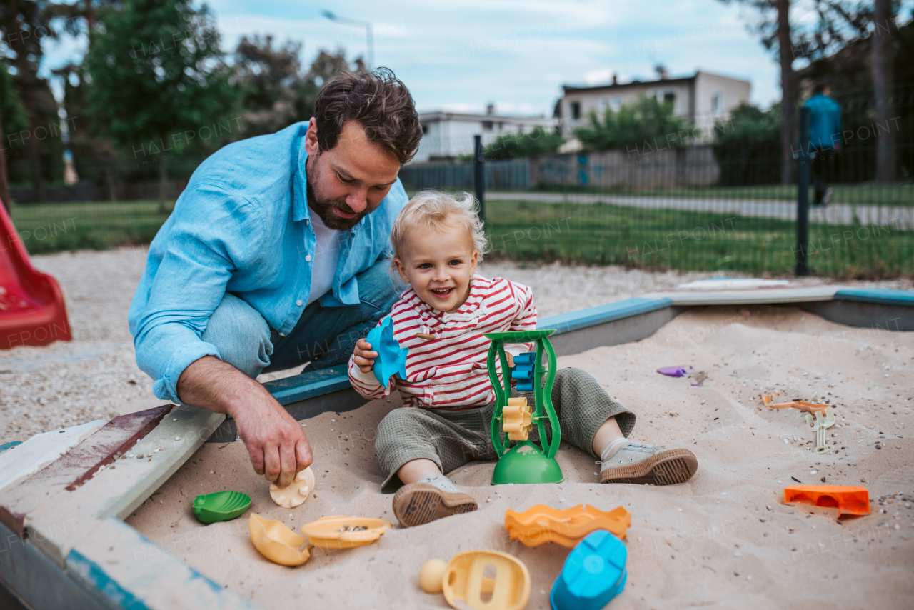 Father and little toddler son playing in the sandbox on playground, weekend fun.