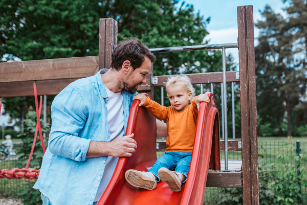 Toddler on a slide with dad's support. Father and son having fun on playground.