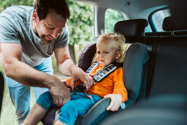 Toddler frowns while dad buckles him into car seat, leaving playground.