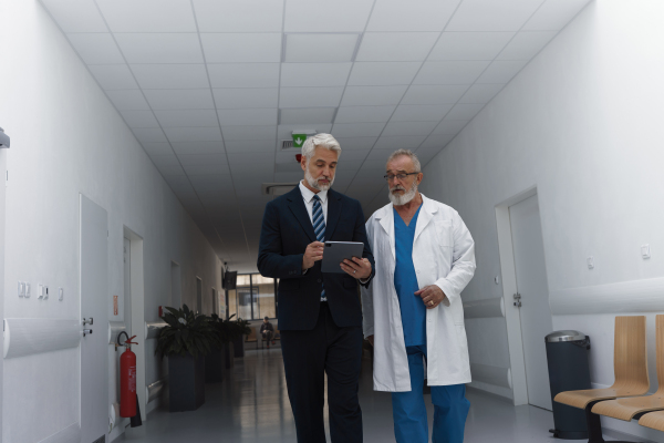 Manager walking with doctor down the hallway, showing him document in tablet. Hospital setting.