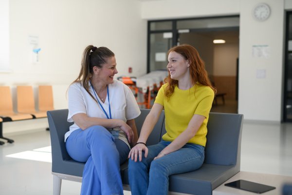 Nurse talking with teenage girl patient in modern hospital waiting room.