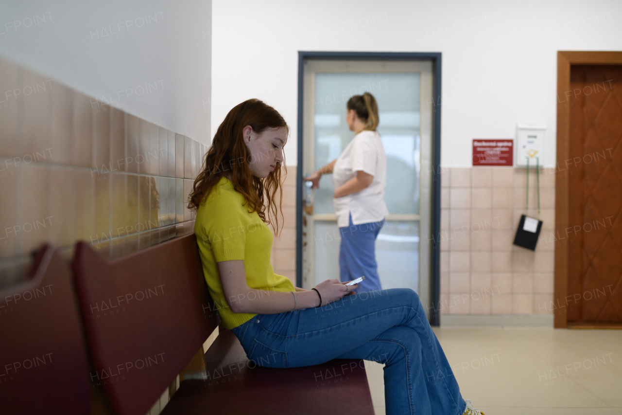 Teenage girl waiting alone in hospital waiting area and scrolling on phone.