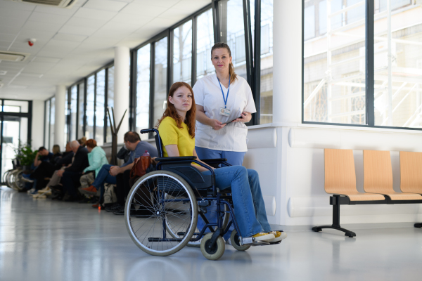 Nurse with young female patient in wheelchair at hospital, modern clinic.
