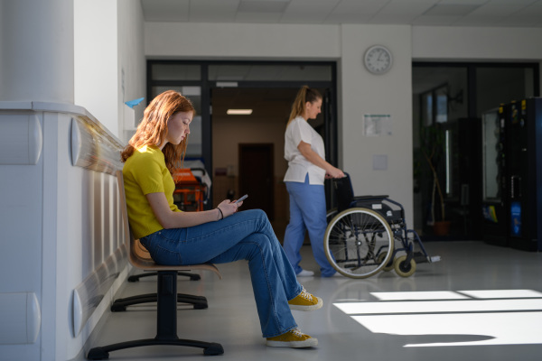 Teenage girl waiting alone in hospital waiting area and scrolling on phone.
