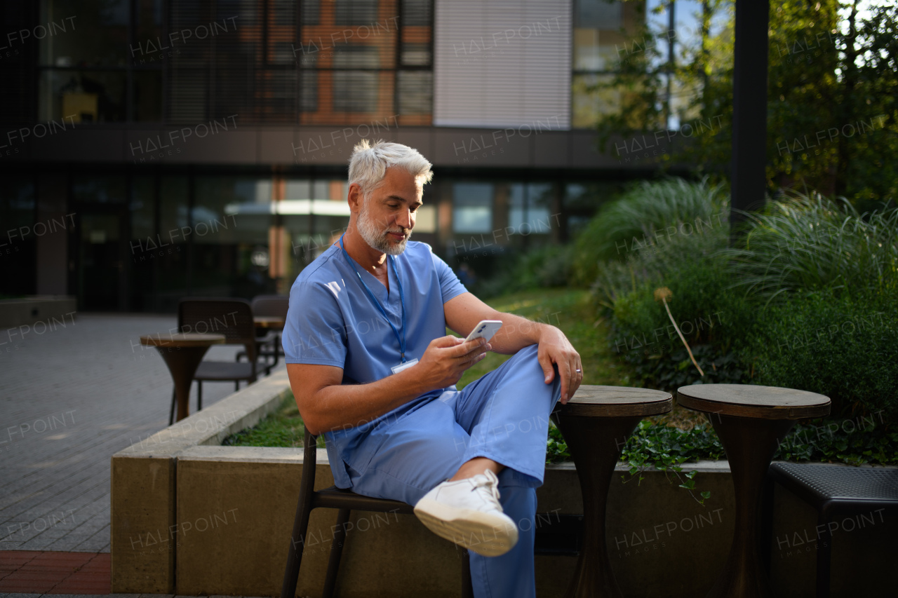 Healthcare worker in scrubs sitting outside hospital and checking phone during break.