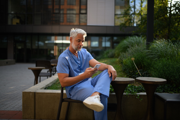 Healthcare worker in scrubs sitting outside hospital and checking phone during break.