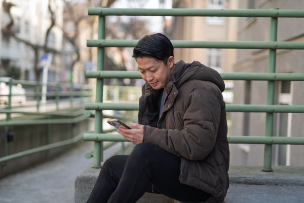 Young Asian man sitting alone on street ledge scrolling on smartphone.