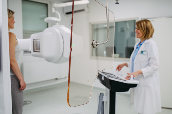 Female patient undergoing dynamic X-ray scan using tilting fluoroscopy wall unit, radiographic table with wall stand.