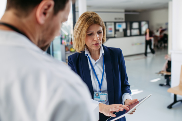 Female medical sales representative presenting medication to doctor, standing in hospital hallway.
