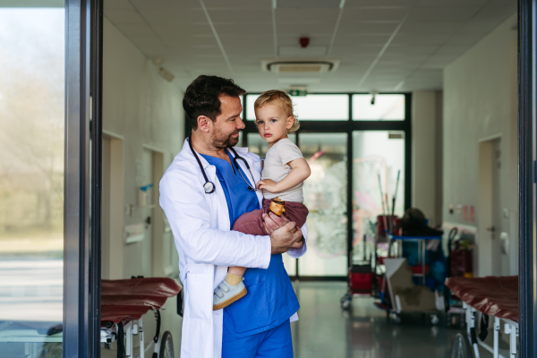 Handsome doctor holding young pediatric patient in arms. Healthcare worker as working father.