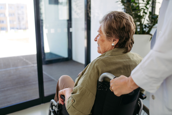 Healthcare worker pushing elderly patient in wheelchair across hospital hallway, Senior health care.