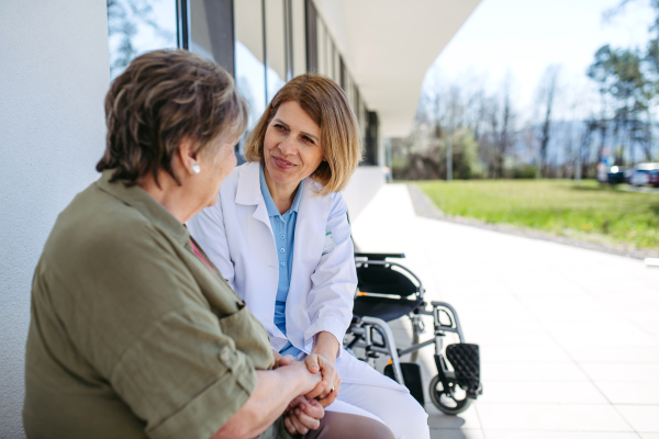 Doctor and elderly patient sitting in front of hospital building and talking. Senior health care.