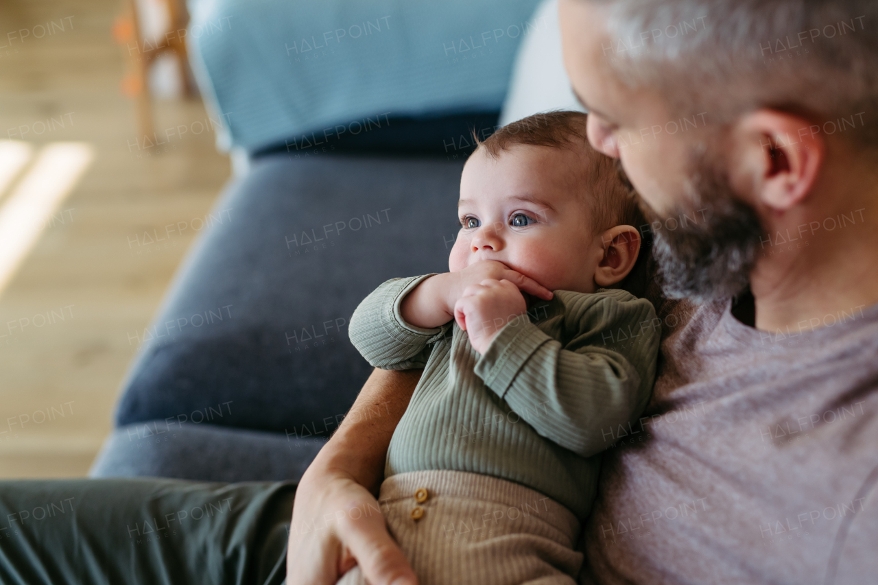 Father holding little baby boy in arms, sitting with him on sofa.