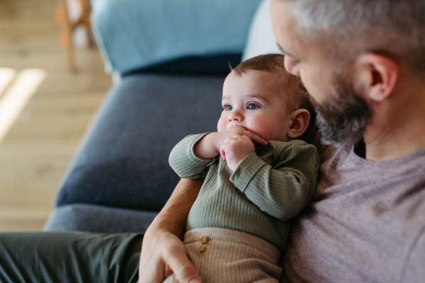 Father holding little baby boy in arms, sitting with him on sofa.