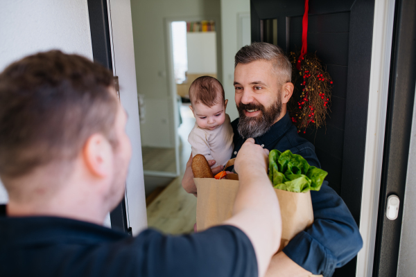 Father holding a baby in his arms receiving a grocery delivery. Online grocery delivery service.