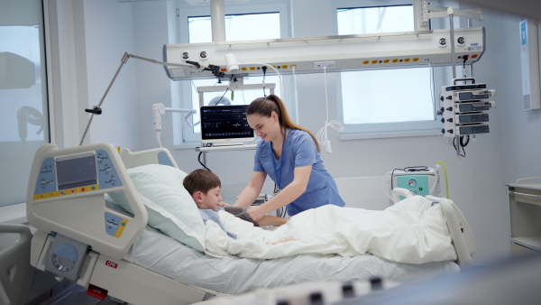 Female nurse caring for young boy patient lying in hospital bed.