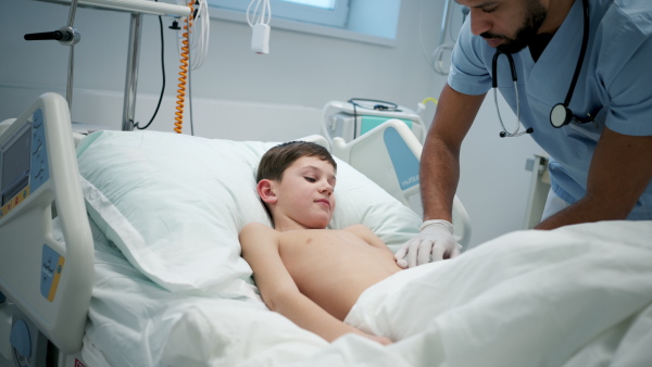 Male doctor examining young boy patient lying in hospital bed.
