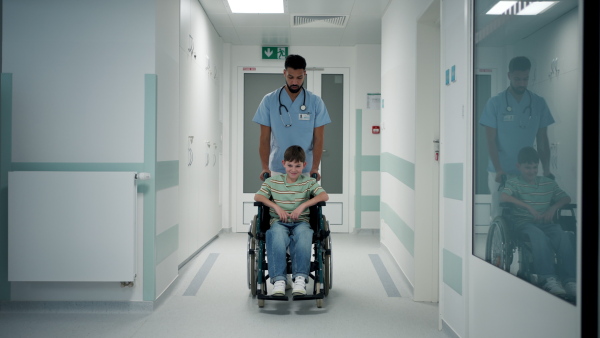 Doctor pushing boy in wheelchair through hospital corridor.