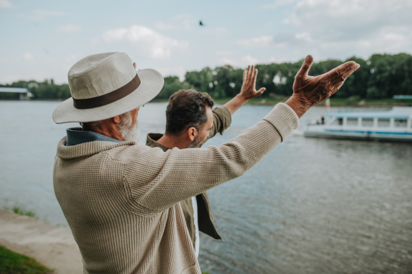 Father and adult son standing on the riverbank and waving at a passing boat. Fathers day concept.