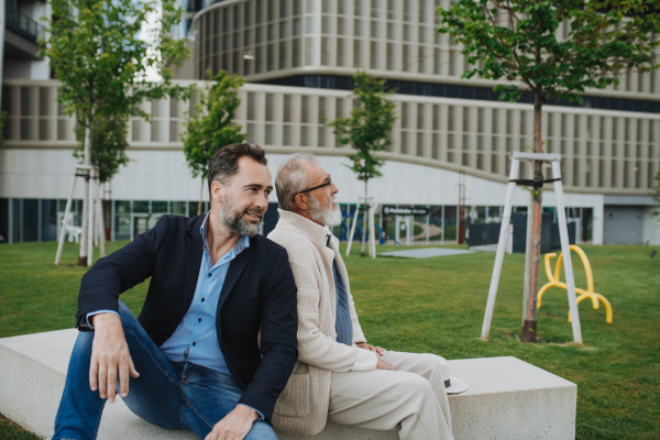 Father and adult son sitting on bench in park, sharing peaceful moment. Fathers day concept.