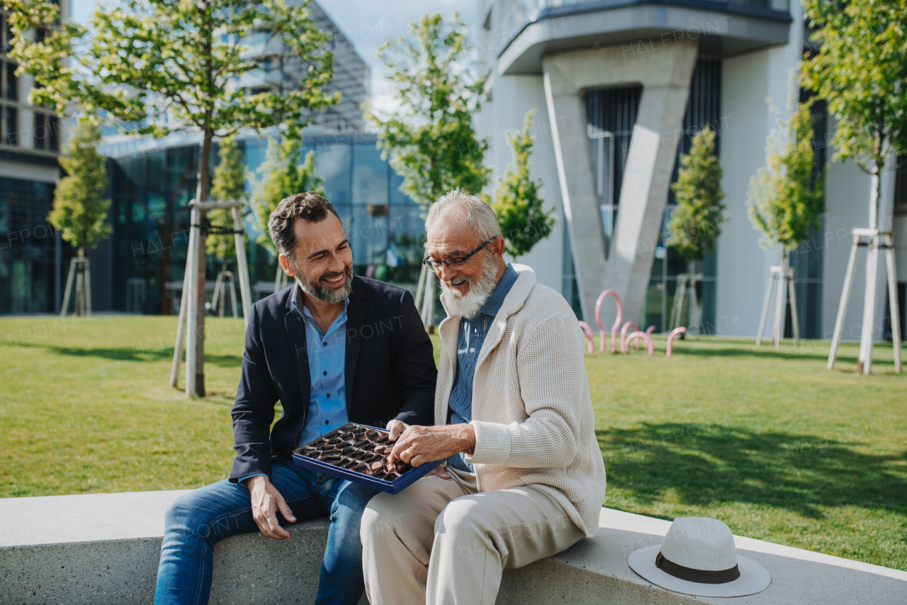 Senior dad and adult son sharing a box of sweets, chocolates on park bench. Fathers day.