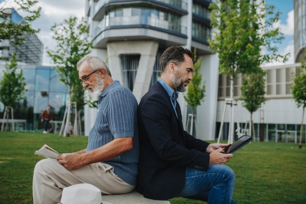 Father and adult son sitting back to back in city park, reading newspaper. Fathers day concept.