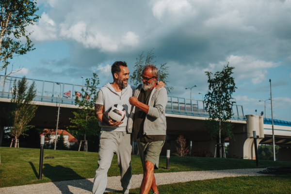 Sporty father and adult son walking home after football match in park. Fathers day concept.
