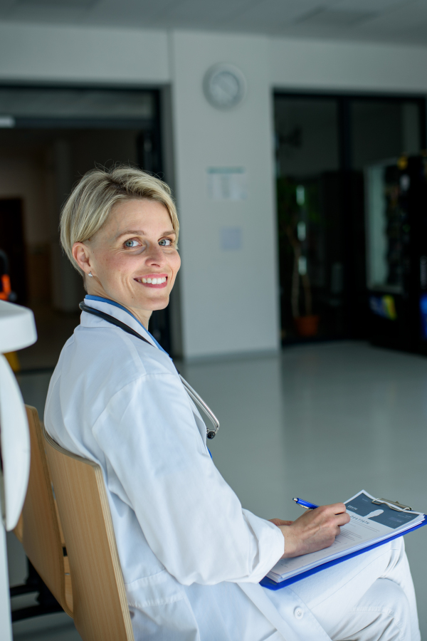 Young female doctor with short hair in sitting on bench in hospital corridor, smiling, looking at camera.