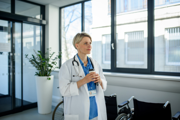Young female doctor taking a break, drinking coffee in hospital corridor.