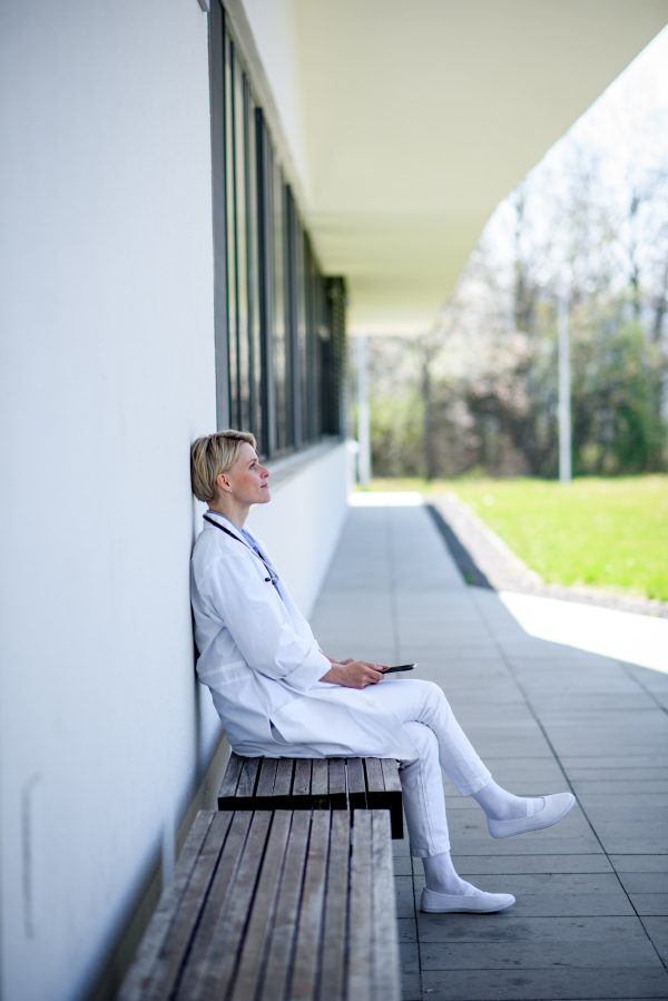 Young female doctor taking a break, sitting on bench in front of hospital building.