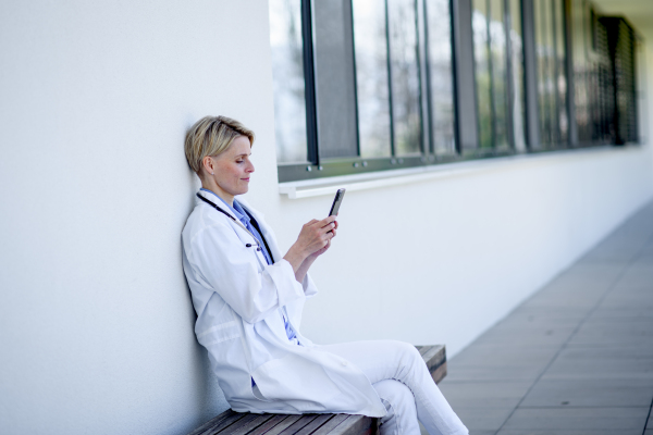 Young female doctor taking a break, sitting on bench in front of hospital building.