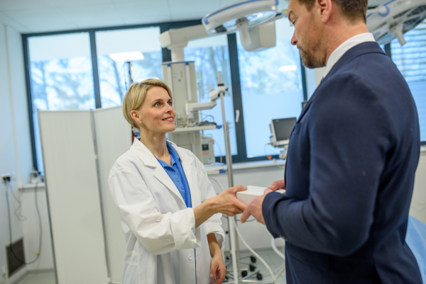 Pharmaceutical sales representative handing a sample box of new medication to female doctor in clinical setting.