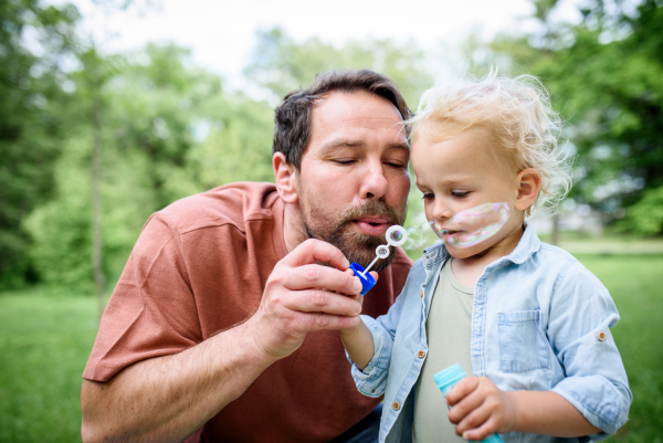 Father and son blowing soap bubbles in park. Toddler watching bubbles with amazement.