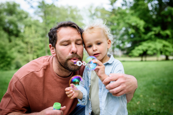 Father and son blowing soap bubbles in park. Toddler watching bubbles with amazement.