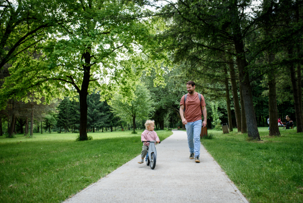 Toddler riding balance bike with dad cheering behind and running behind him.