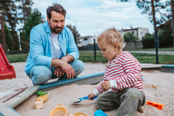 Father and little toddler son playing in the sandbox on playground, weekend fun.