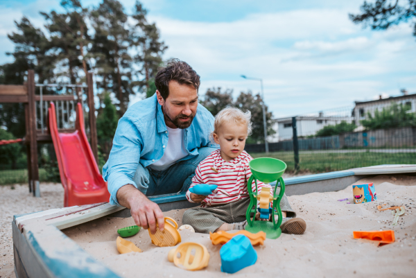 Father and little toddler son playing in the sandbox on playground, weekend fun.