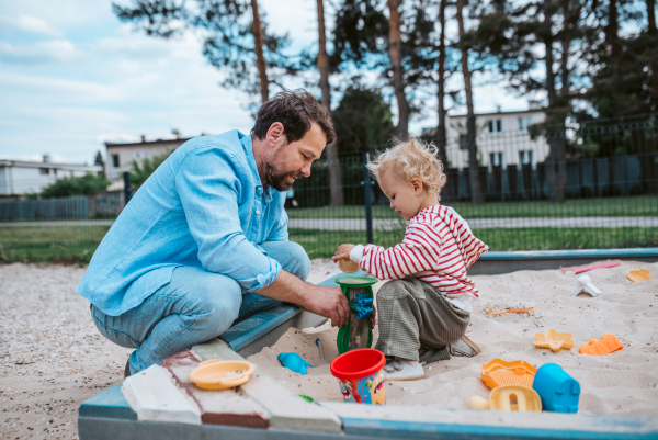 Father and little toddler son playing in the sandbox on playground, weekend fun.