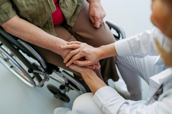 Doctor holding hands with elderly woman in wheelchair during comforting talk.